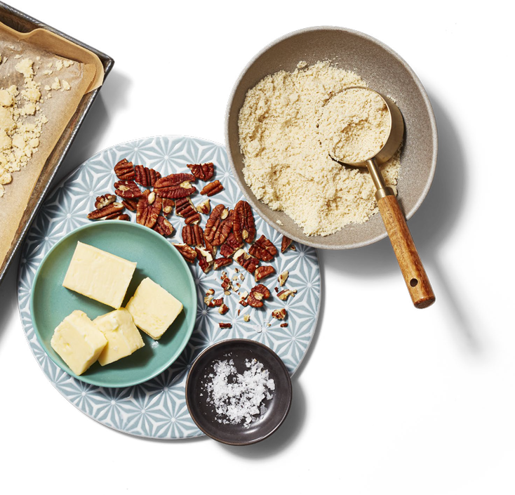 Un plat de cuisson carré tapissé de papier parchemin et un bol de service sont remplis des ingrédients pour préparer des carrés au dulce de leche à côté d'une assiette de noix de Grenoble hachées, de bâtonnets de beurre et de flocons de sel.