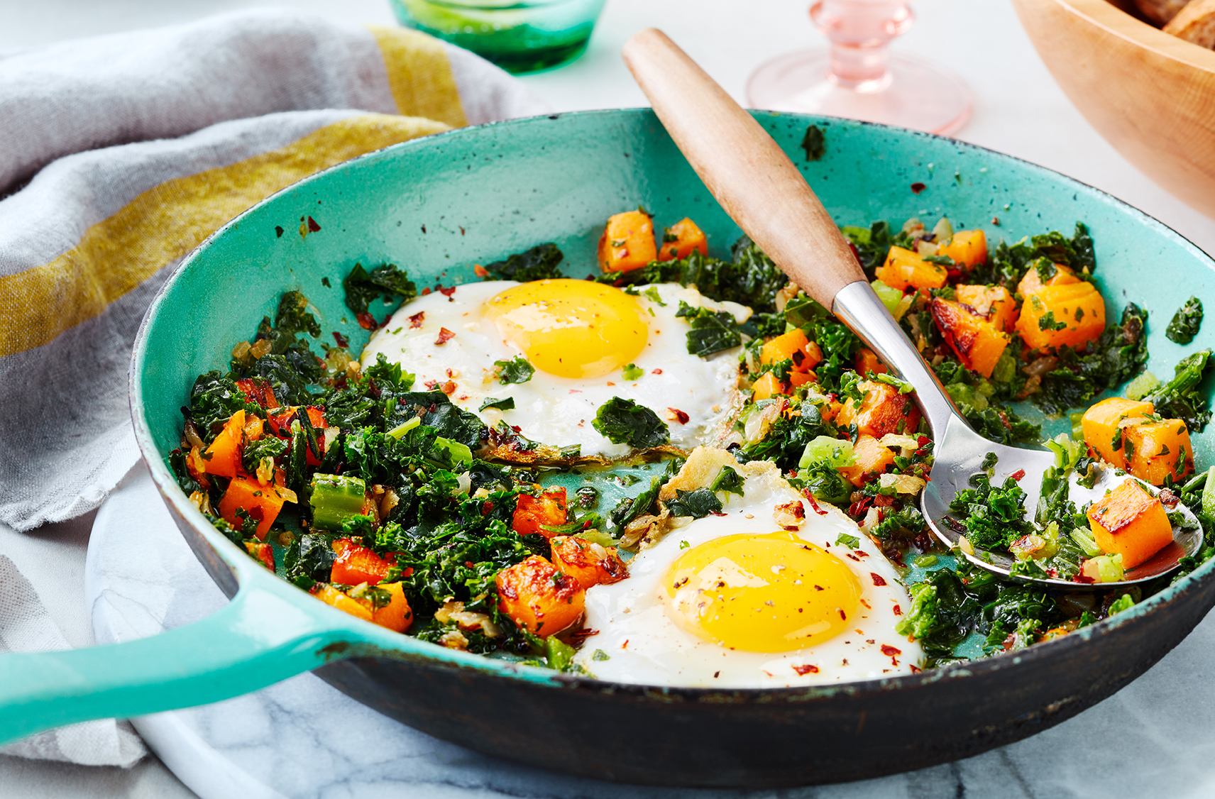 A spoon serves fried eggs over sautéed greens and cubed squash from a pan
