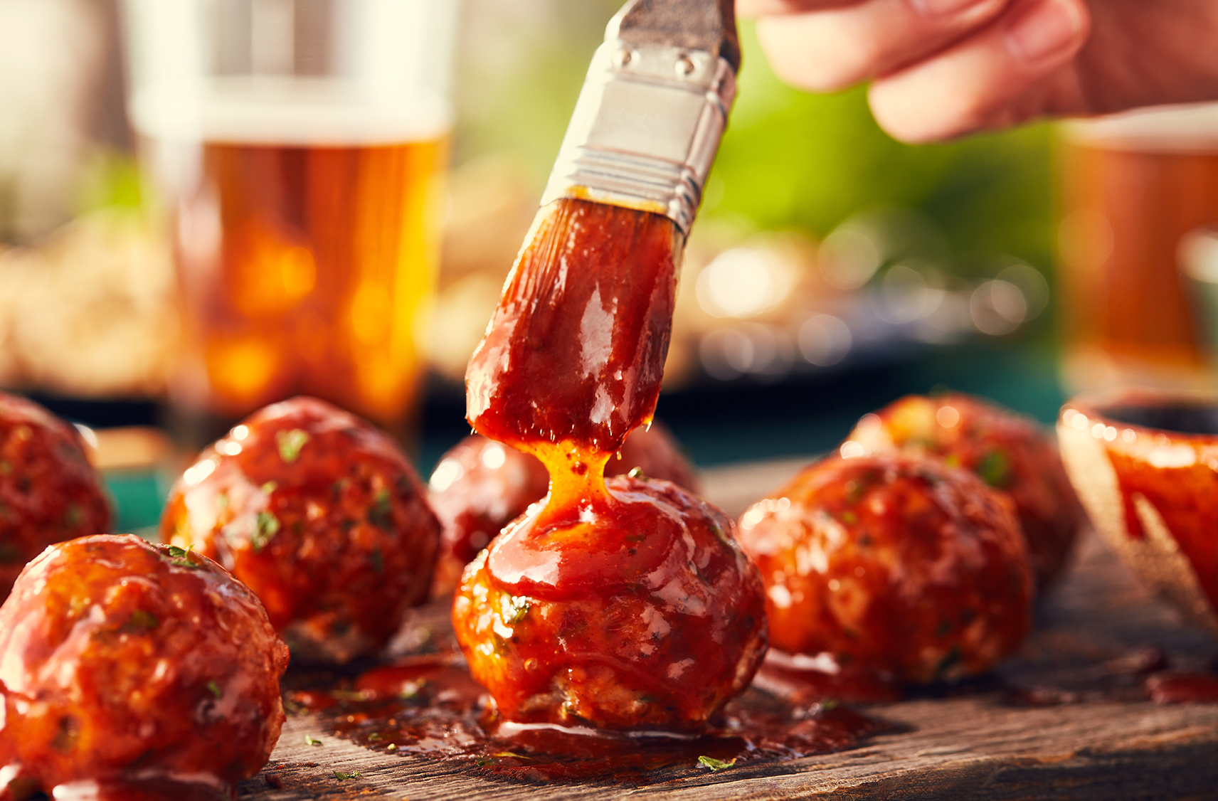 A pastry brush coats meatballs with sauce, arranged on a wooden board