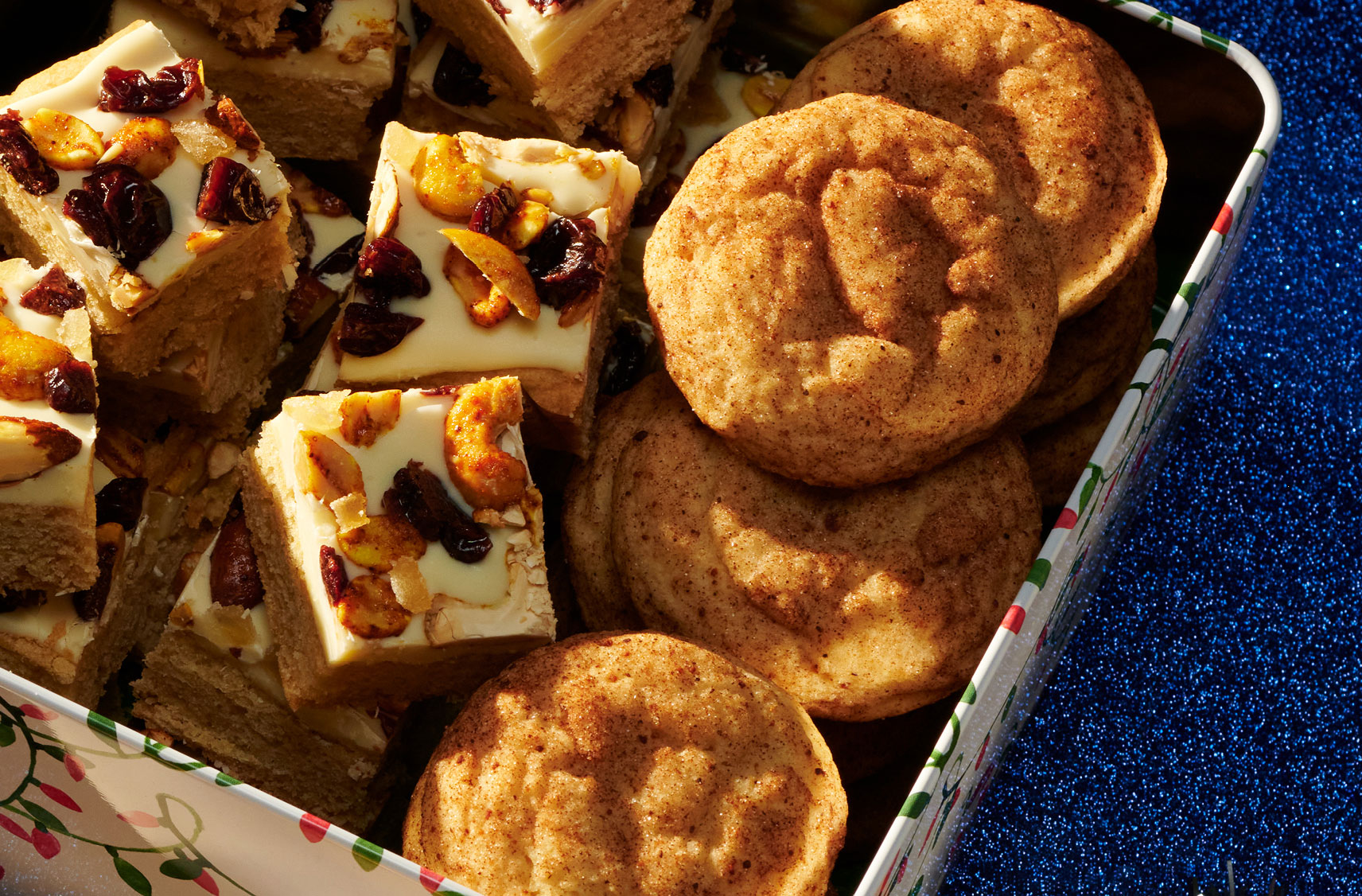Une boîte en métal remplie de biscuits, dont des snickerdoodles au lait de poule et des carrés au chocolat blanc et au curcuma sur une table.