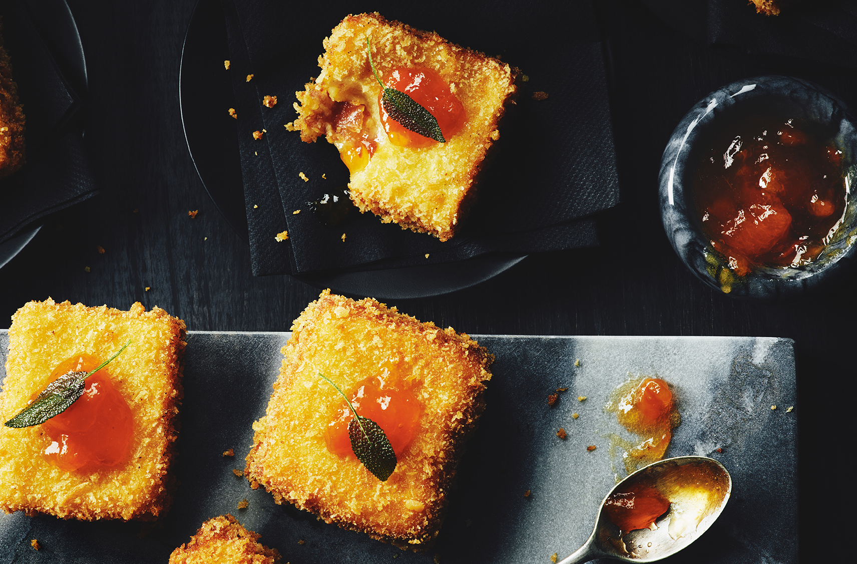 3 pieces of crispy cheese mini sandwiches topped with fried sage and a dollop of apricot riesling jelly.  One of the mini sandwiches has a bite taken out of and placed on a small black plate