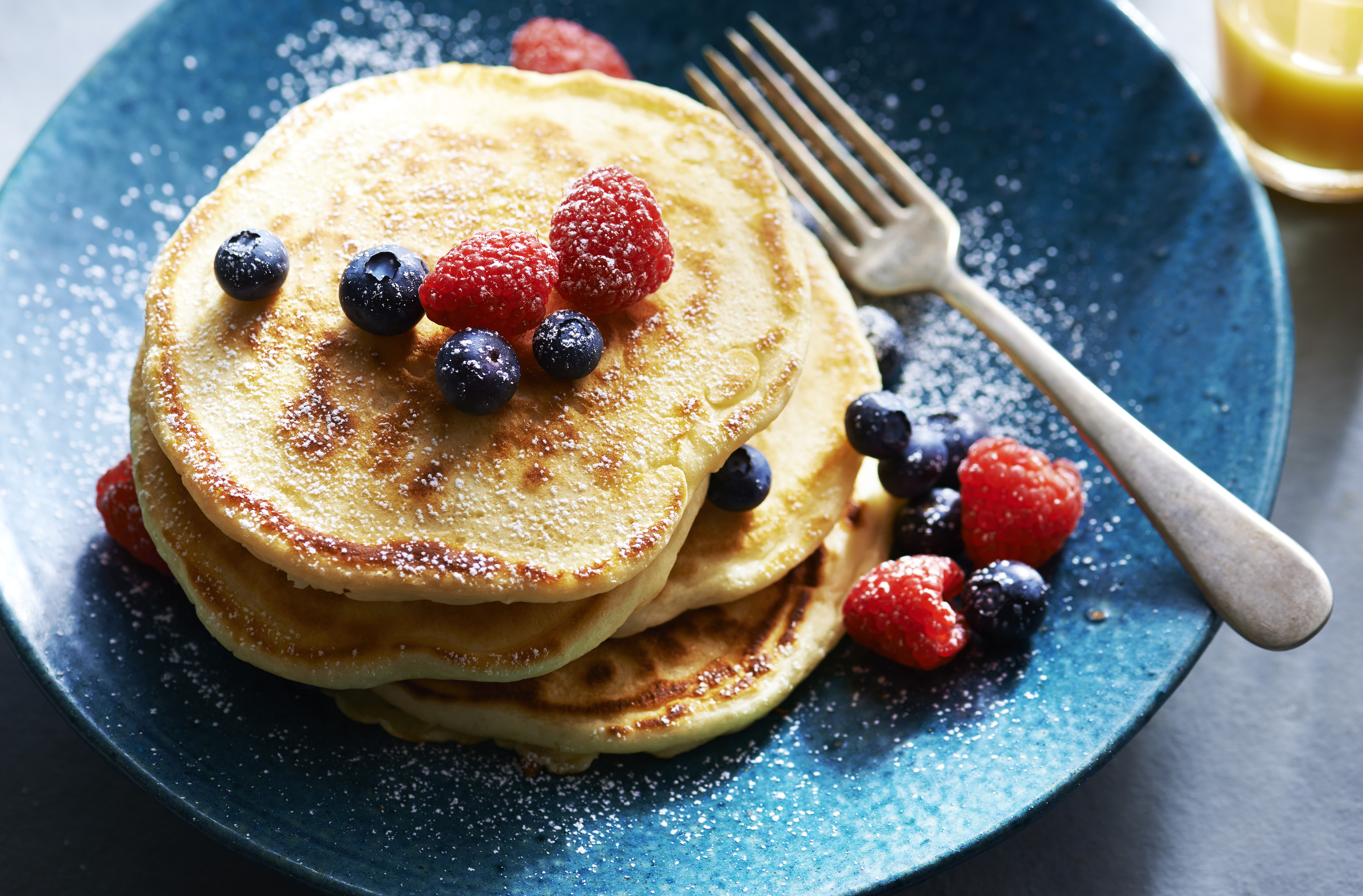 4 Kefir pancakes stacked on a plate.  Topped with blueberries and raspberries and dusted with powdered sugar