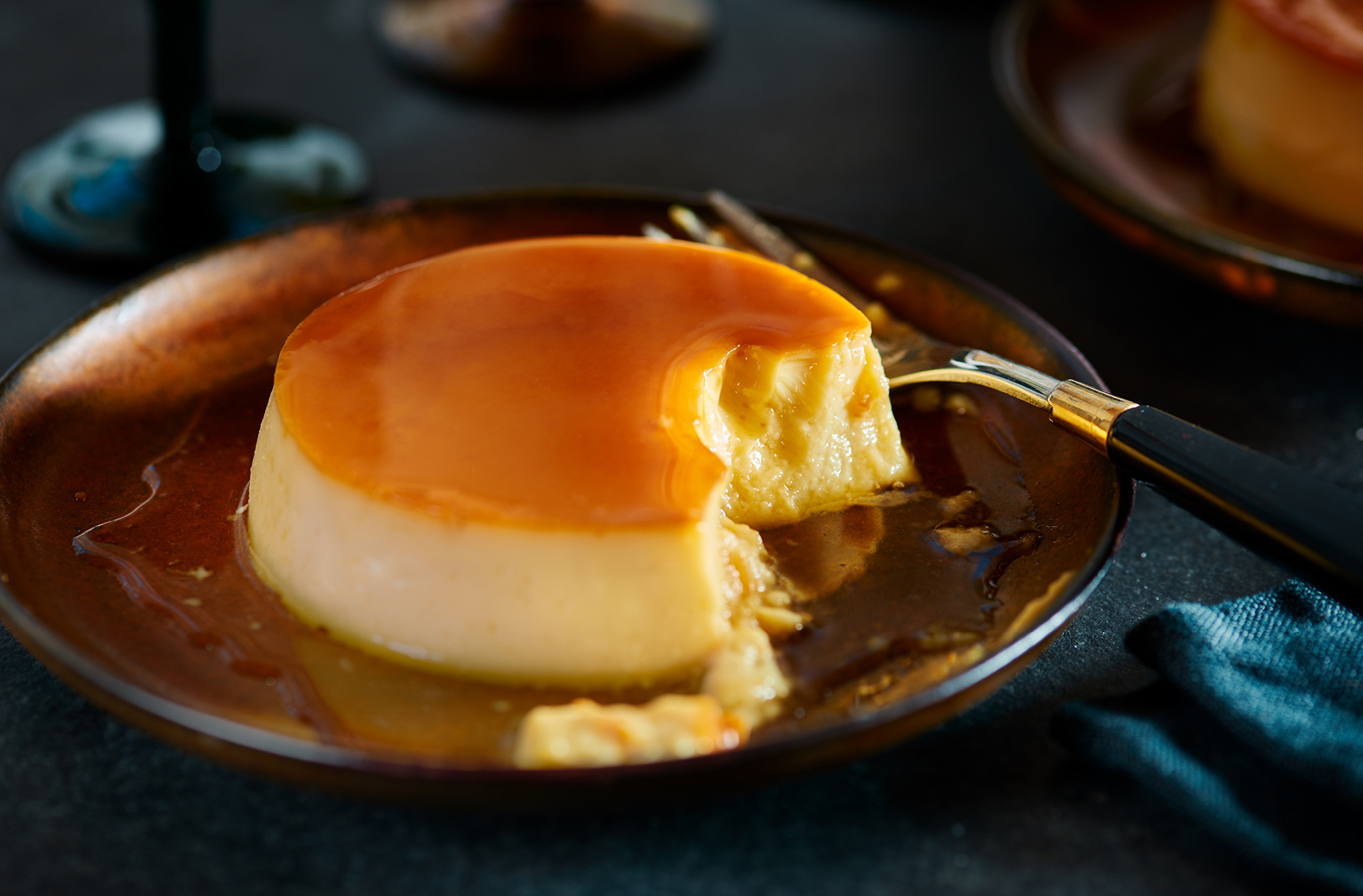 A crème caramel dessert on a plate next to a fork with 1 bite missing