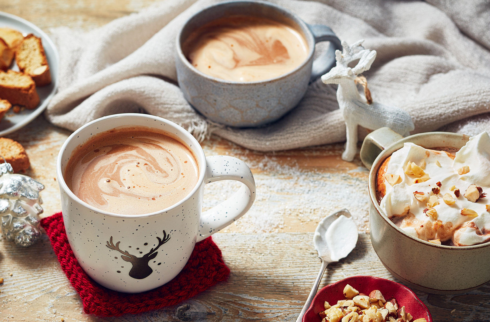 Three chocolate drinks in mugs, with one topped with whipped cream and nuts sitting next to a mini reindeer.	