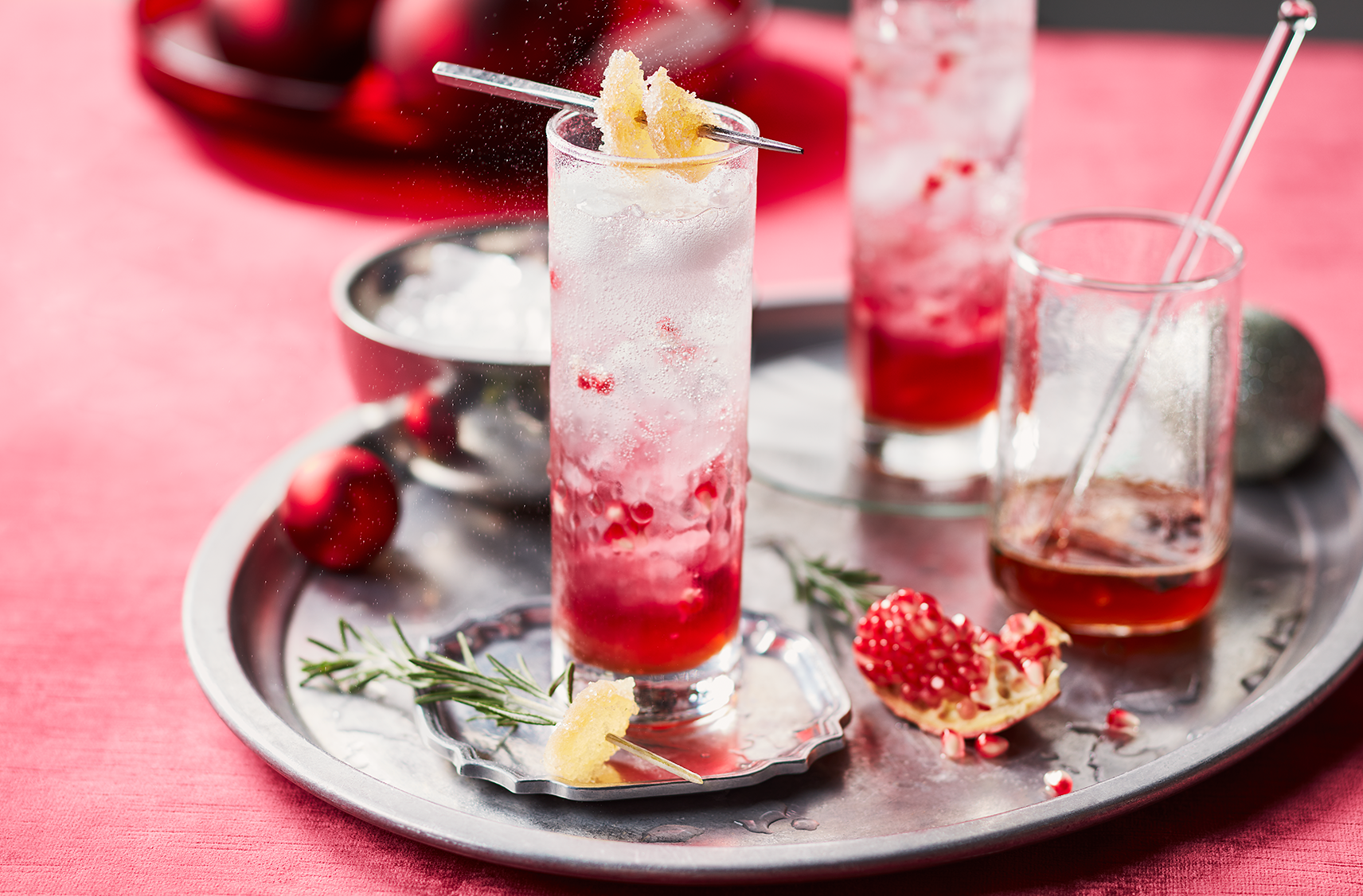 2 glasses of sparkling ginger pomegrante mocktail beverages on top of a siver serving tray with ice and syrup on the sides