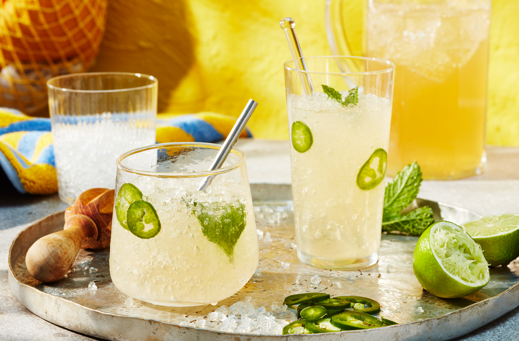 Two glasses of a lemon and ginger cocktail garnished with lime, mint leaves and jalapeno slices served on a large tray 