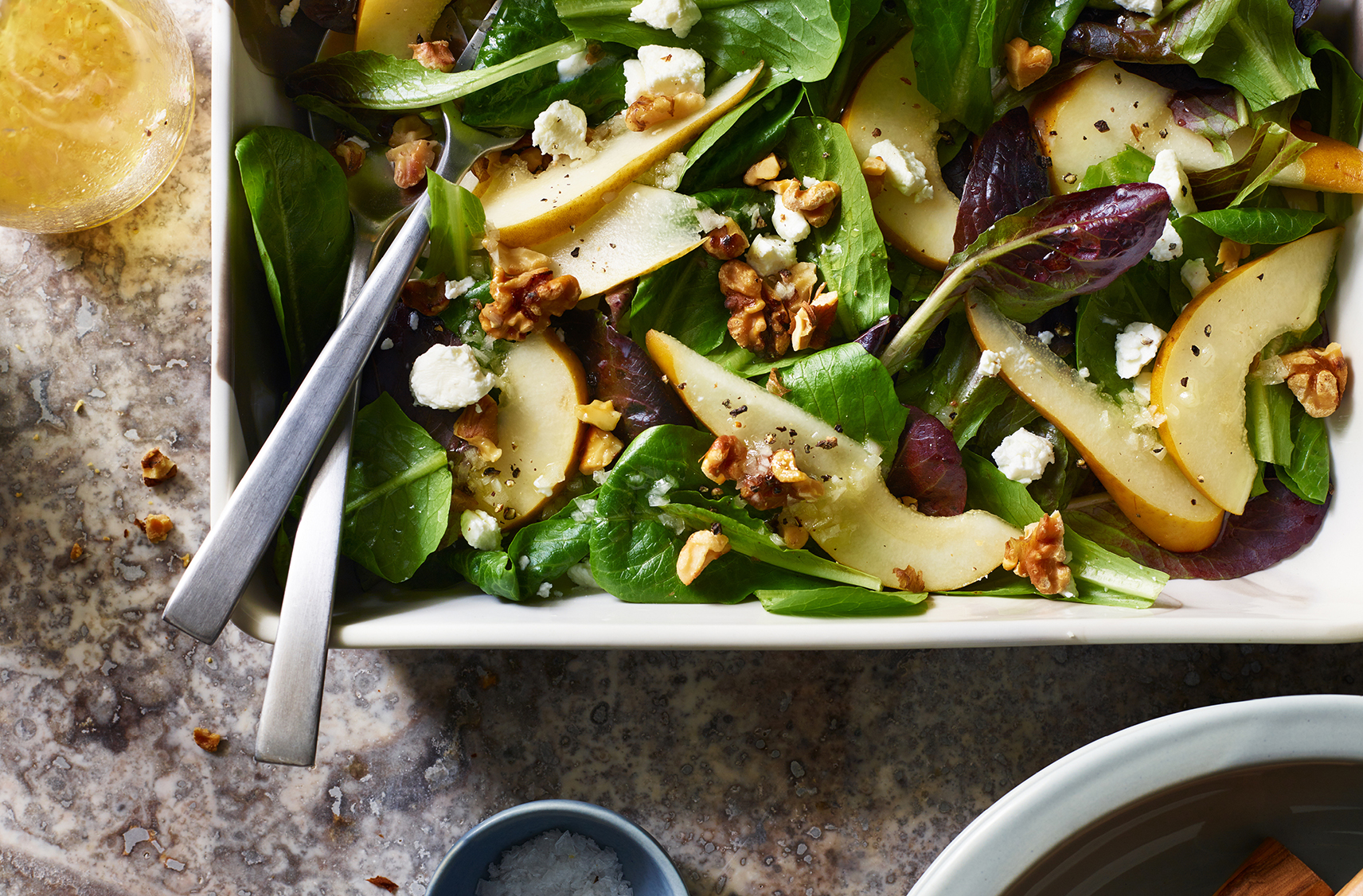 A salad with wild greens, pear slices and crushed pieces of walnuts on top in a serving dish with 2 utensils
