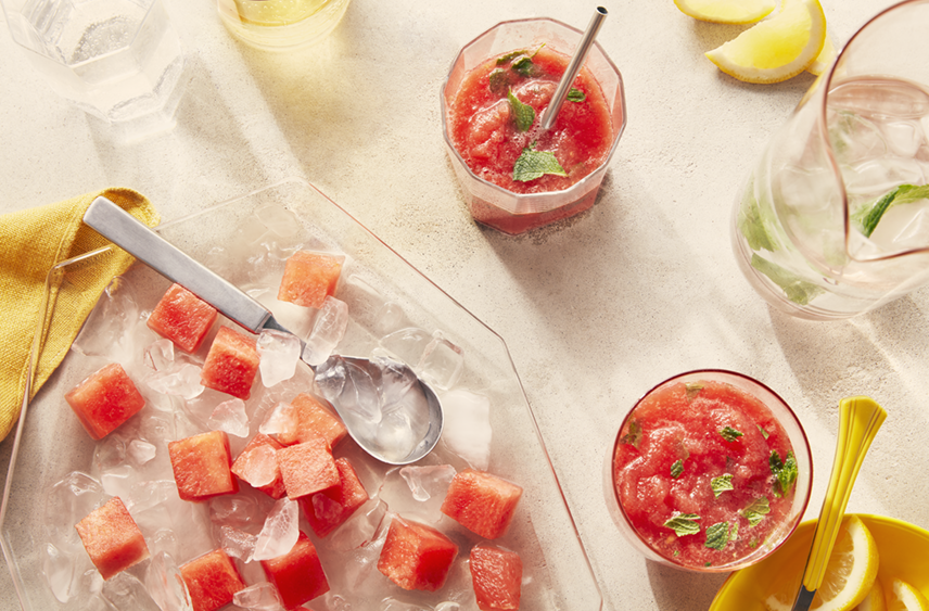 A bowl of PC Frozen Watermelon Chunks with some chunks being added to a glass of Frozen Watermelon Mint Lemonade  beside the bowl

