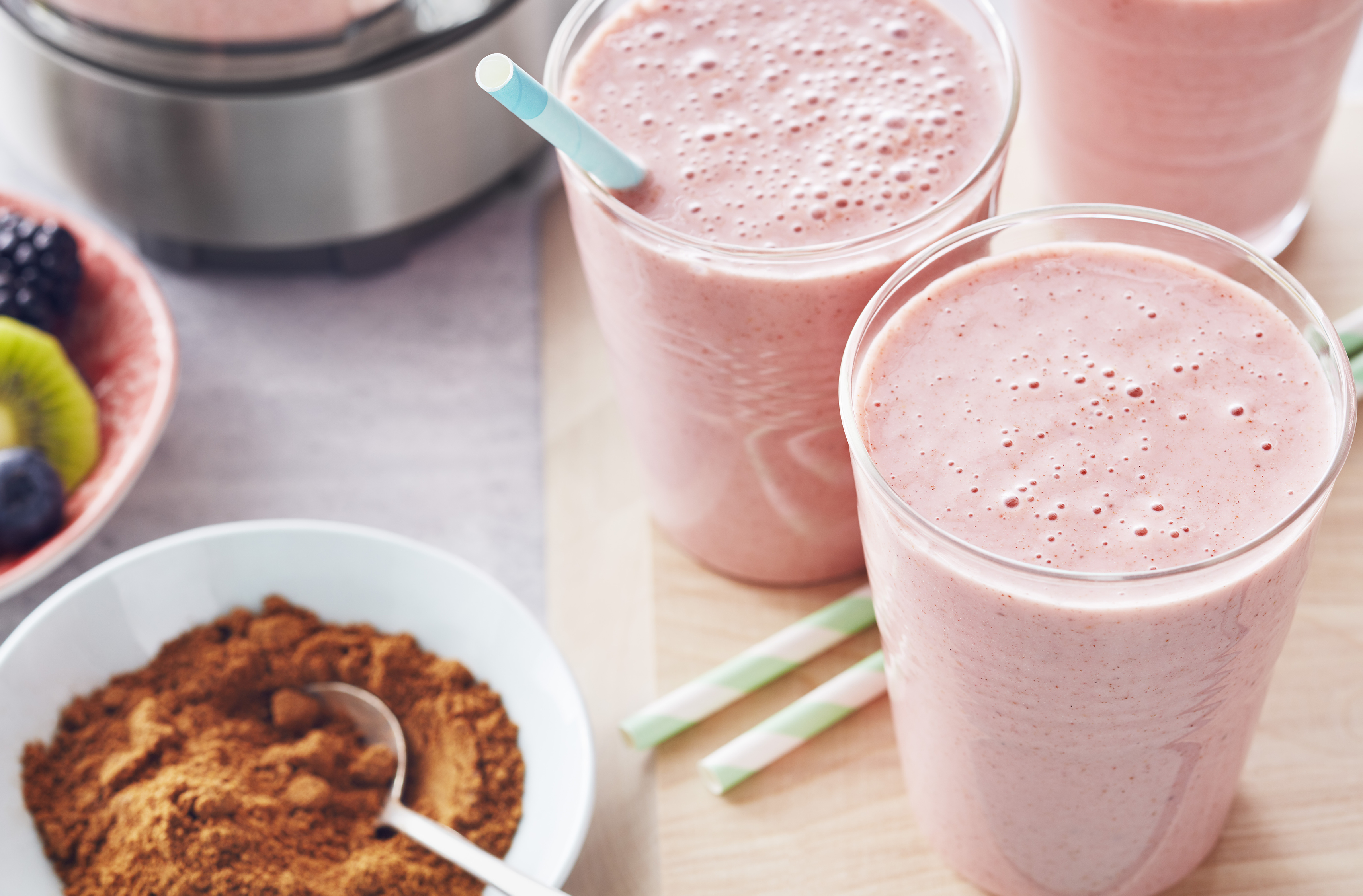 Pink smoothies with straws stand beside fresh fruit and a bowl of spice
