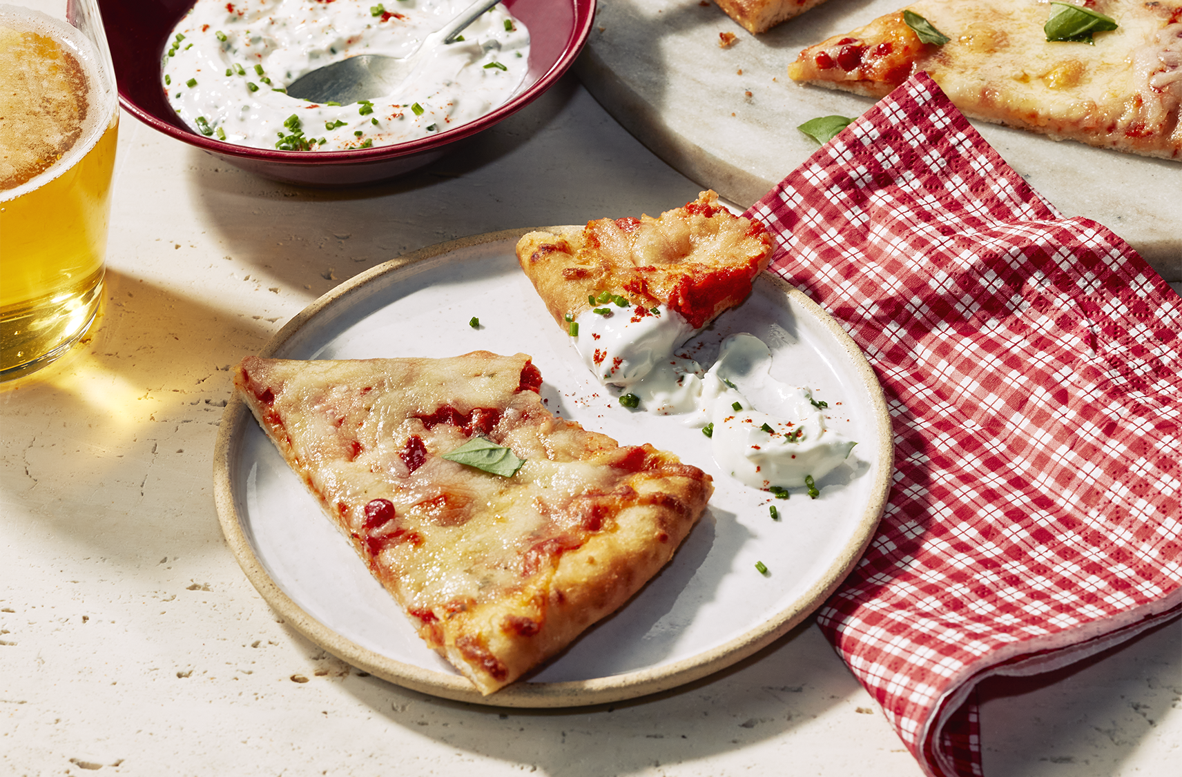 A slice or margherita pizza on a plate with a portion removed and dipped in a herb and garlic dipping sauce. The bowl of dipping sauce and a beverage sit beside the pizza.