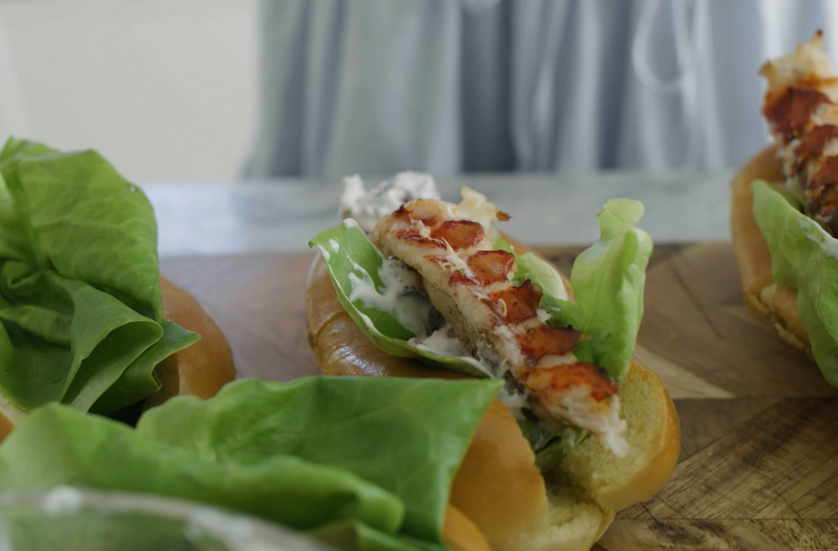 Des pains à hot dog garnis de laitue fraîche sur une table à côté d'autres pains déjà préparés avec une brochette de queue de homard et de la sauce crémeuse.