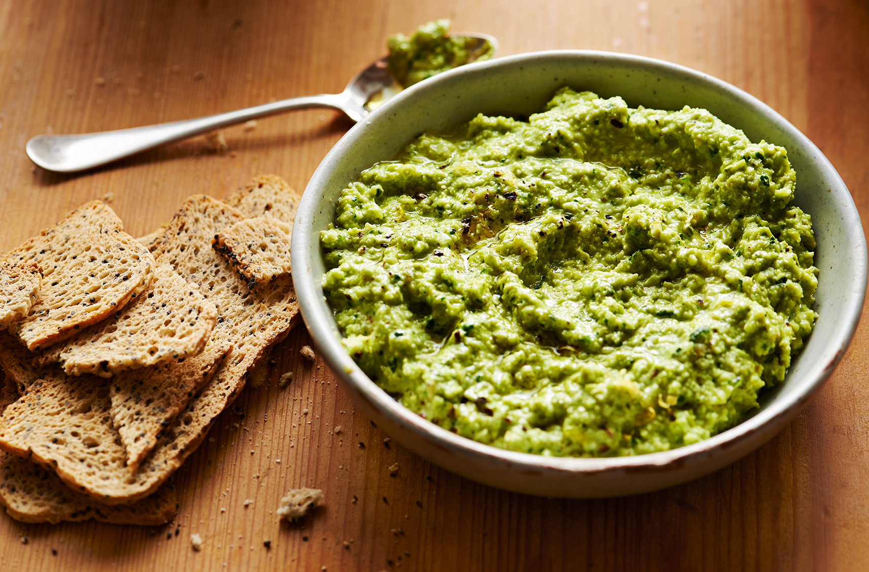 Du hummus vert servi dans un petit bol gris avec des biscottes pour tremper