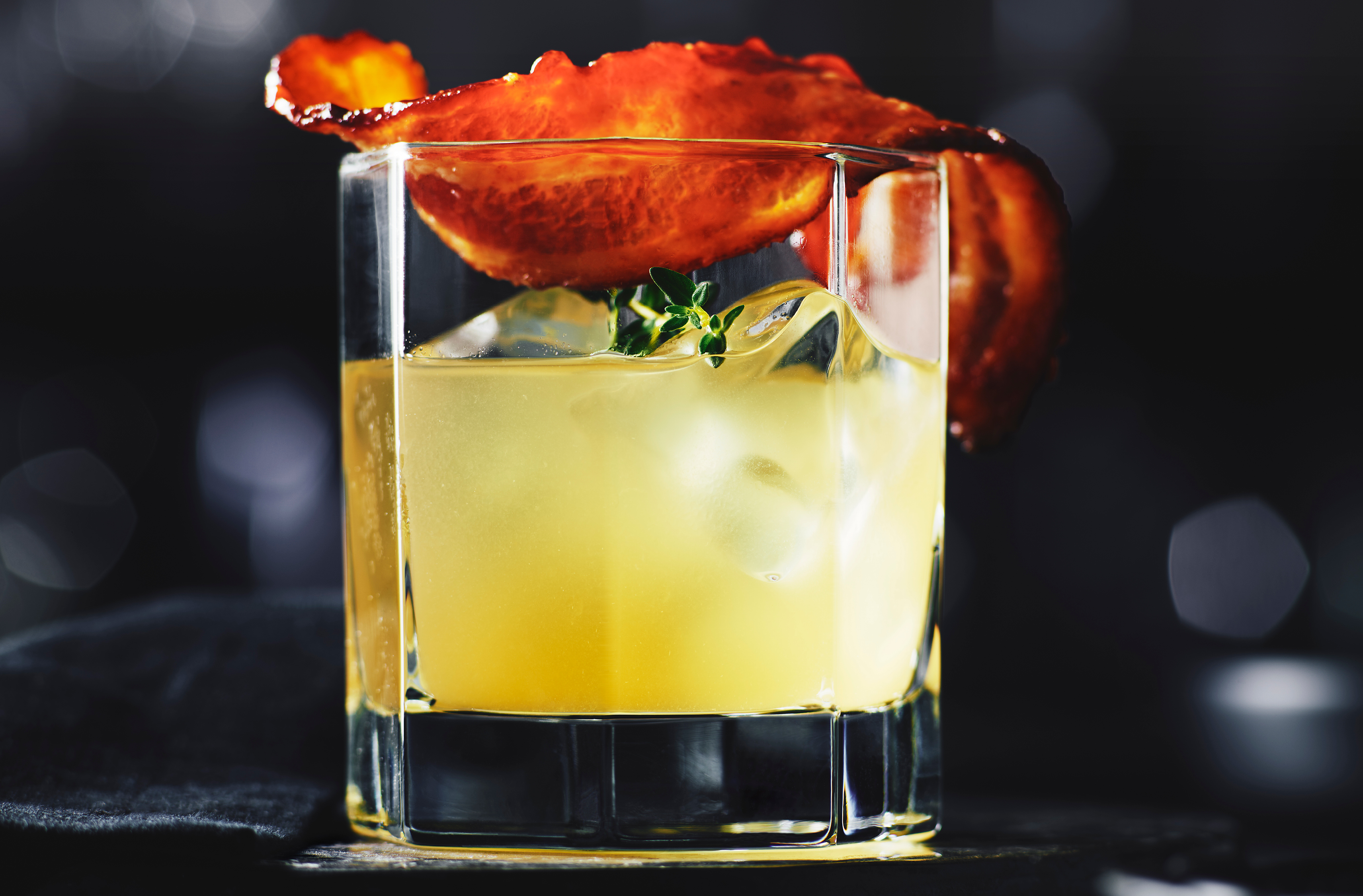 A glass of white peach bourbon shrub garnished with candied bacon
