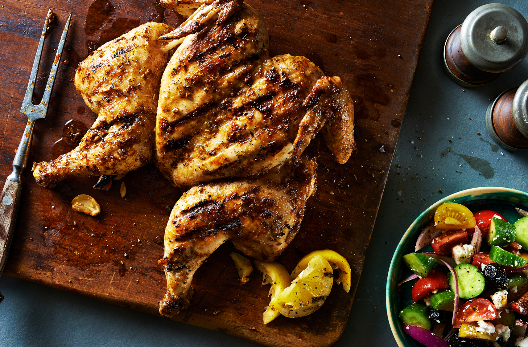 Un poulet à la grecque grillé en crapaudine placé sur une planche à découper en bois et servi avec une salade en à côté