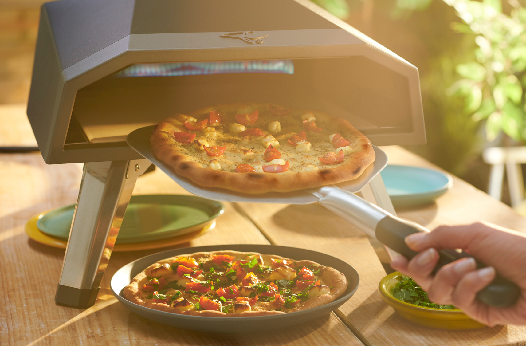 Une Pelle à pizza PC qui sort une pizza garnie de tomates, de fromage halloom et de fines herbes d’un Four à pizza au propane PC.
