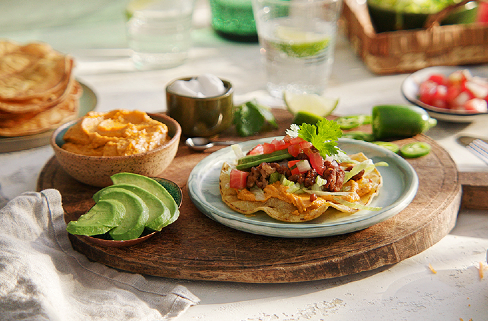A wooden board with a plate holding a dressed plant based tostado with slises of avocaado and a bowl of plant based dip on the side.
