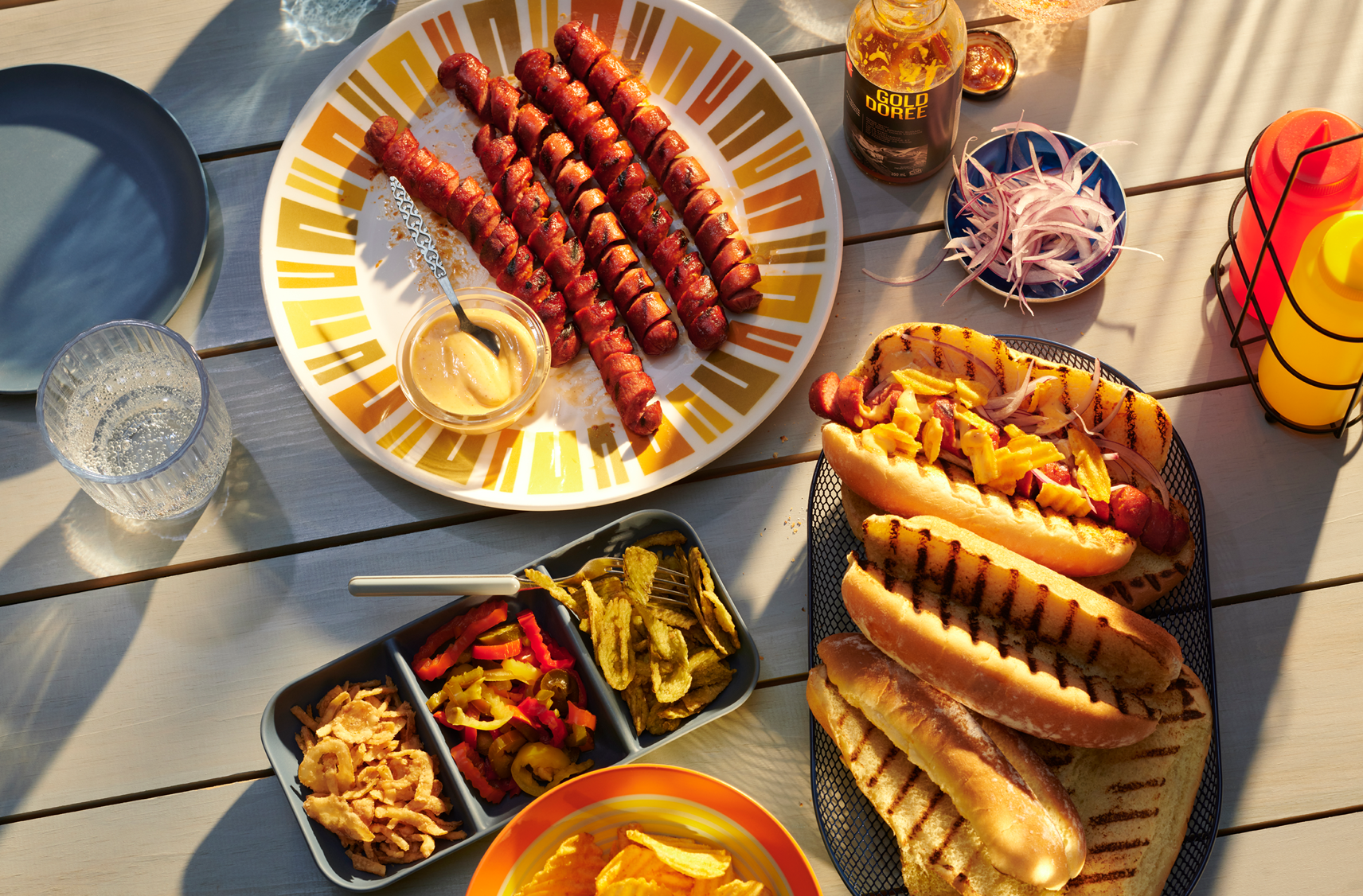 A picnic table with some summer faves including spiralized hot dogs on a dish and grille buns on another and a variety of toppings and condiments.
