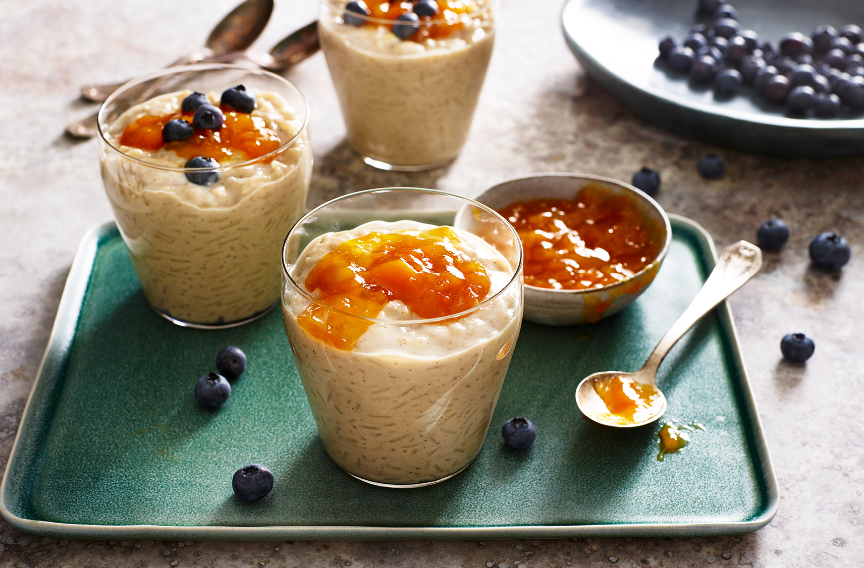 three vanilla rice puddings placed inside dessert glasses topped with apricot jam and fresh blueberries
