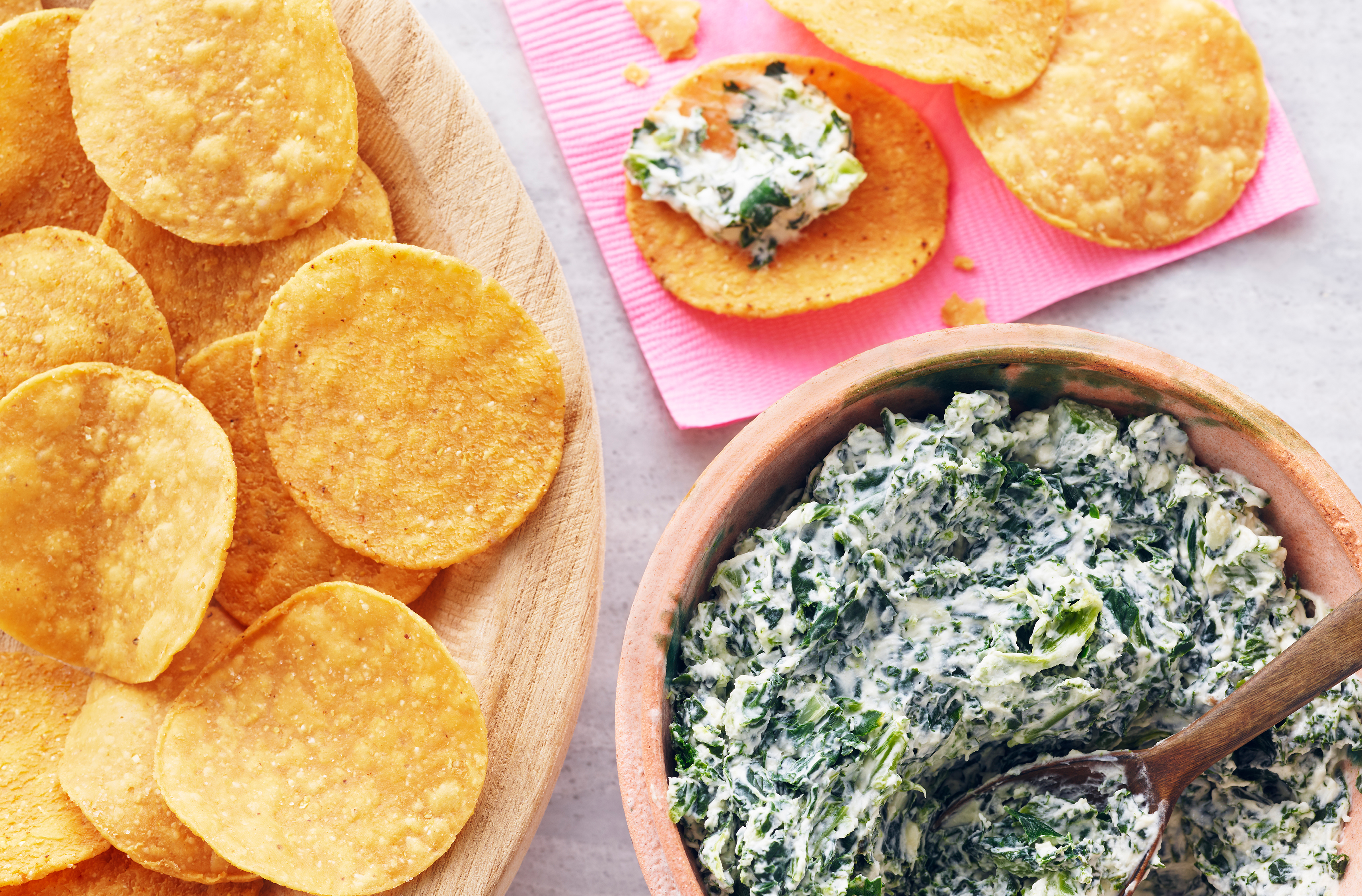 A wooden spoon spreads creamy spinach-and-greens dip onto round nacho chips
