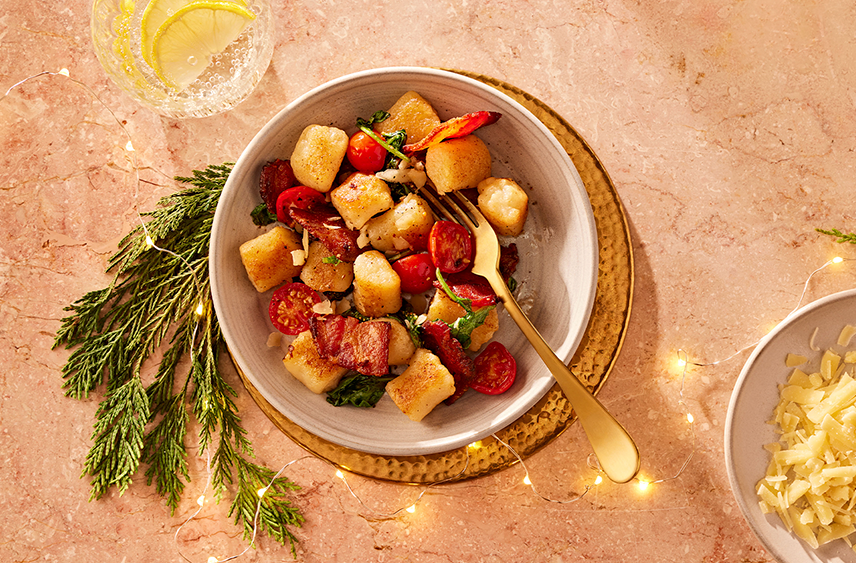 A bowl of PC cauliflower gnocchi sauteed smoky bacon and cherry tomatoes, spinach and parmesan sitting next to a bowl of shredded cheese.