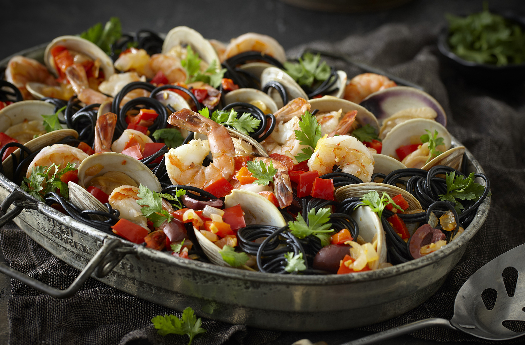 A shallow pot containing black squid ink spaghetti with clams, shrimp & red peppers next to a serving spoon.