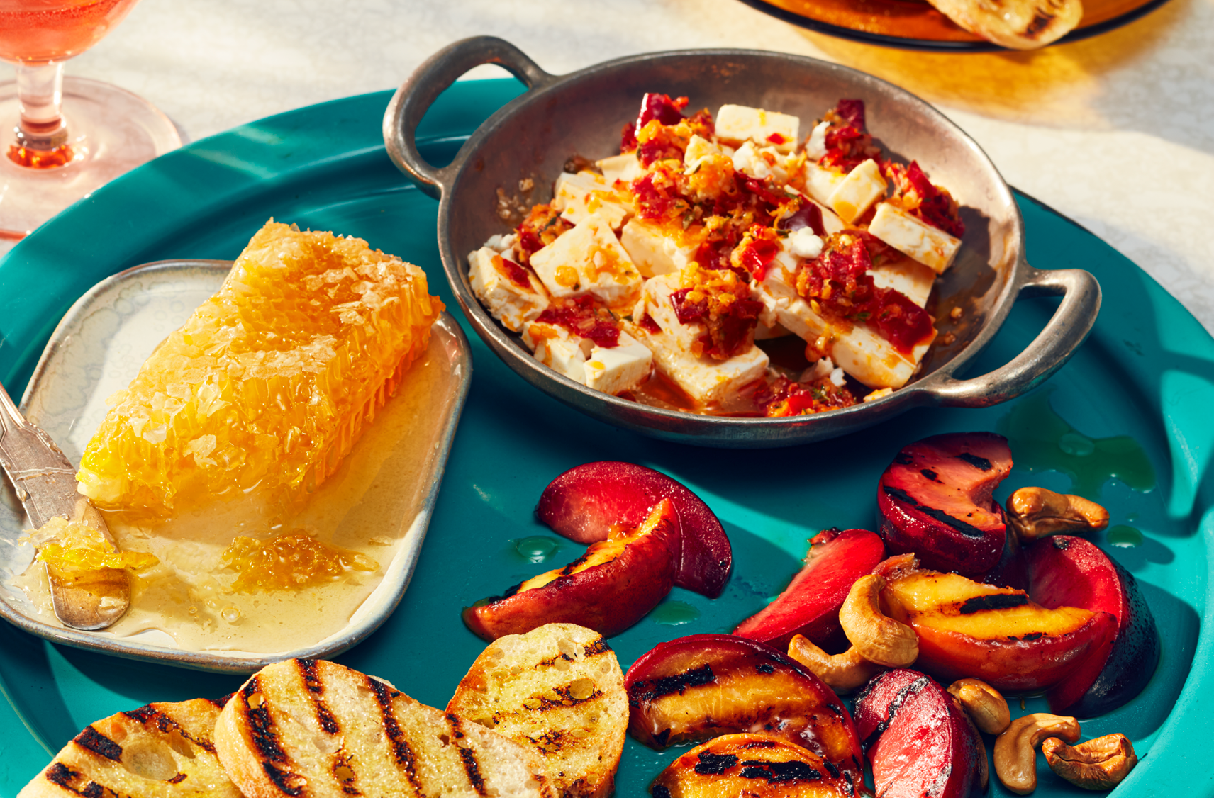 A round tray with the makings of a summer charcuterie including feta with a spicy topper, a large piece of honeycomb with grilled veggies, baguettes and peaches.