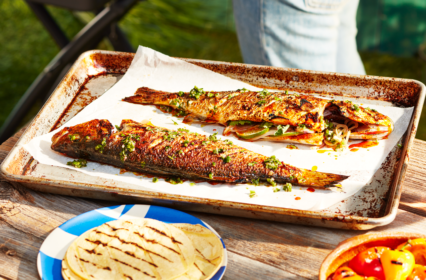 Two pieces of grilled whole fish with topped with Jalapeño Salsa and Sweet Peppers on a sturdy cast-iron grill pan next to a plate of grilled Halloom and another plate of grilled peppers.