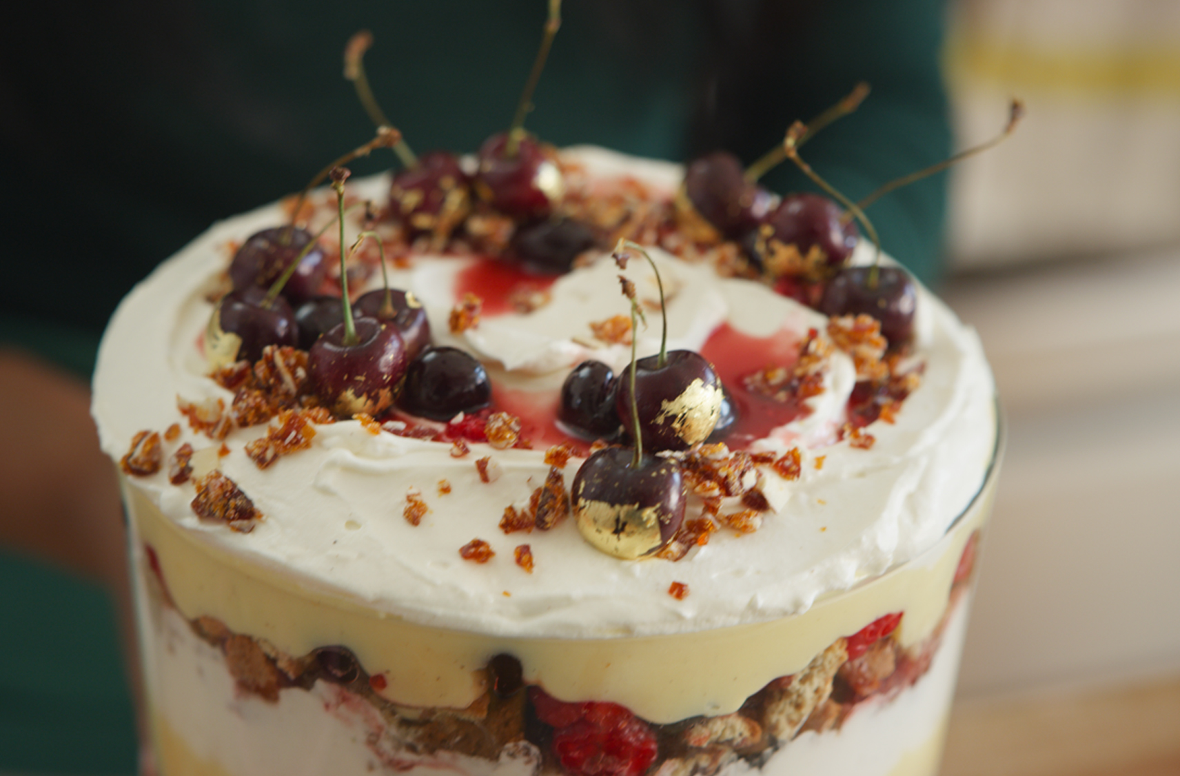 A Cherry Almond Trifle in a glass bowl with layers of whipped cream, berries and custard topped with almond pieces and cherries .