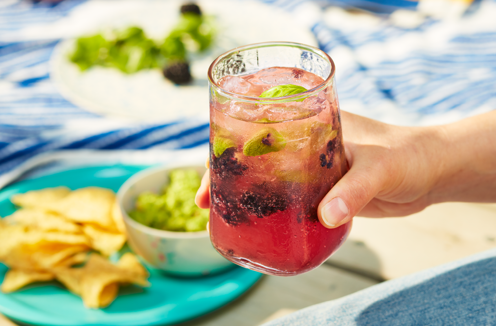 Une main tient un verre rempli de Kéfir d'eau pétillant PC Biologique avec des mûres et du basilic. On peut voir une assiette de tortillas et de guacamole en arrière-plan.