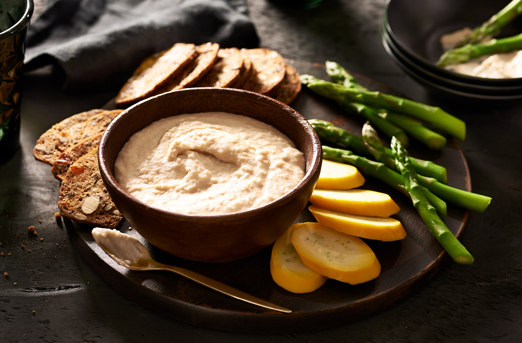 Chipotle and cheddar white bean dip in a small brown bowl.  slices of yellow zuchinni, crackers and asparagus placed around the bowl for dipping