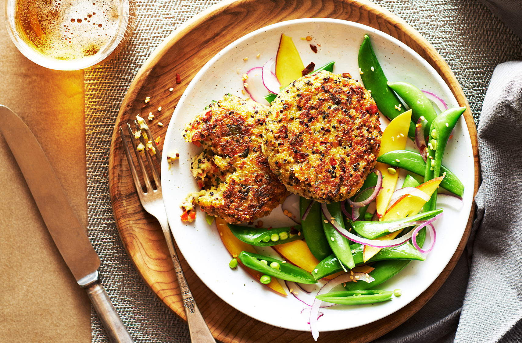 A plate with two thai quinoa patties and some veggies on the side sitting on a bamboo charger palte with a fork knife and beverage on the side