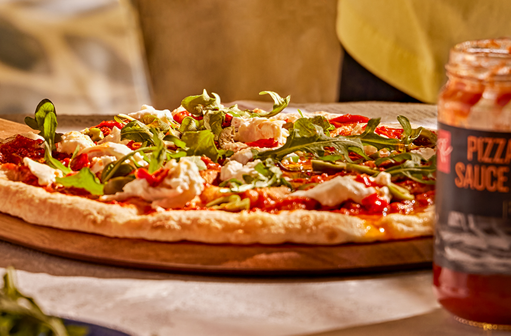 Le Four à Pizza PC est visible au loin, une main arrose d’huile de piment peperoncino une pizza fraîchement cuite, à côté d’un pot de sauce à pizza, d’une salade et de quelques boissons.