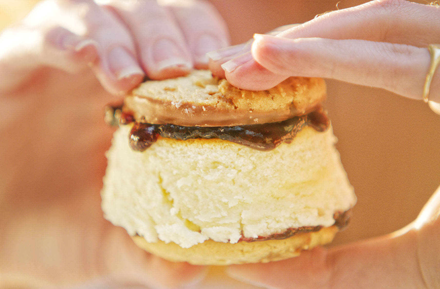 Two hands are holding a cookie sandwich filled with a caramel drizzled cheesecake centre