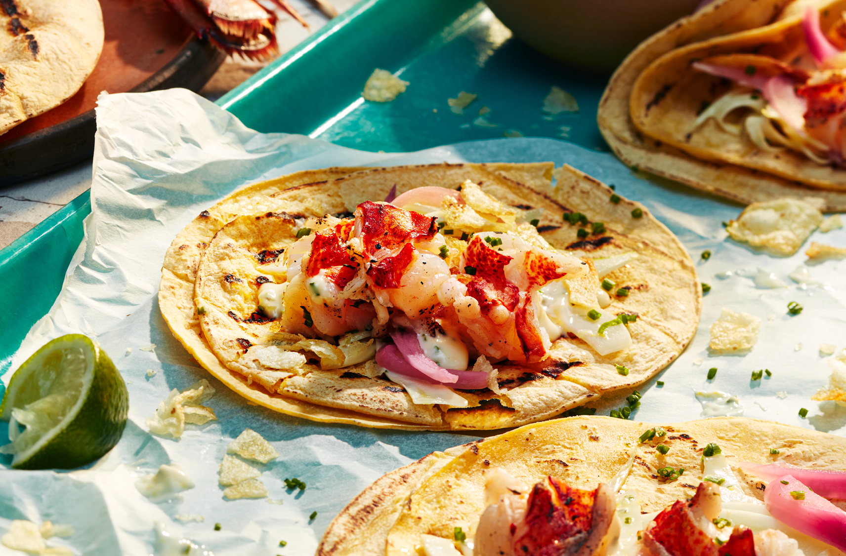 A tray with several tortillas that have PC Lobster Tail Skewers, pickled red onion with a crunchy potato chip topping and herbs.