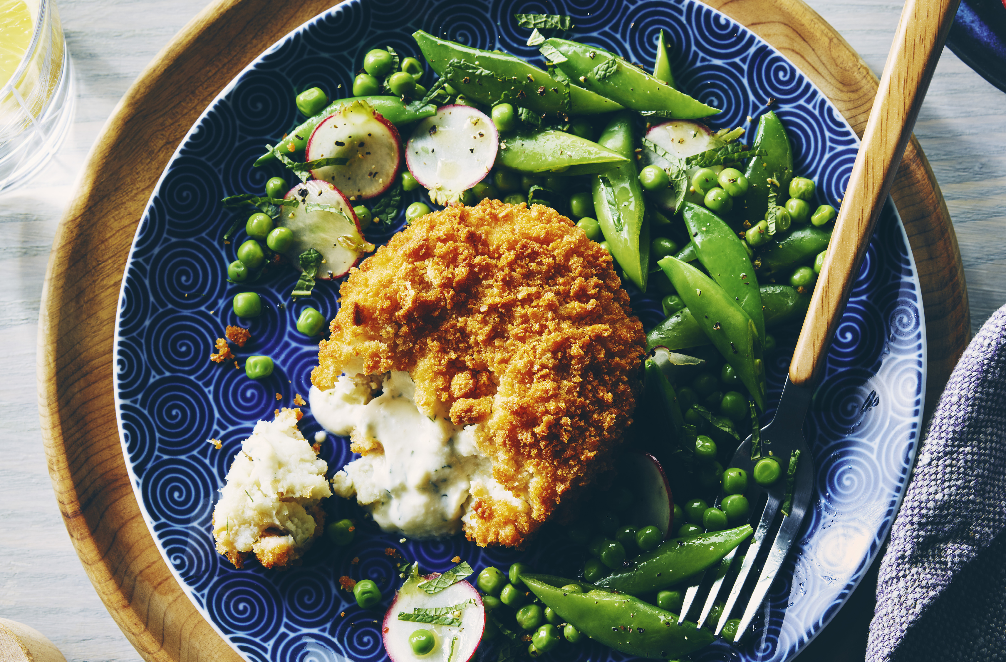 A large breaded cod cake beside snap peas, spring peas and radish slices