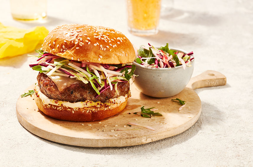 A Smoky Pork Cheeseburger topped with Apple Slaw and served on a cutting board with a serving side of Apple Slaw on the side.