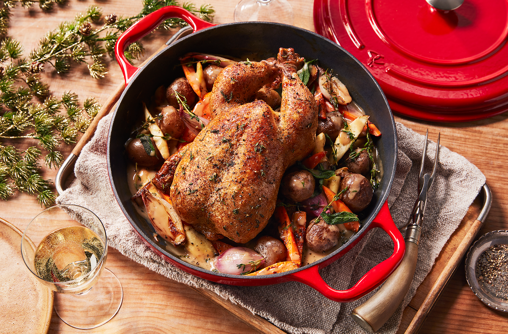 A Dutch stock pot filled with a roasted chicken and root vegetables on a table top with the dutch pot lid off to the side and a sparkling beverage