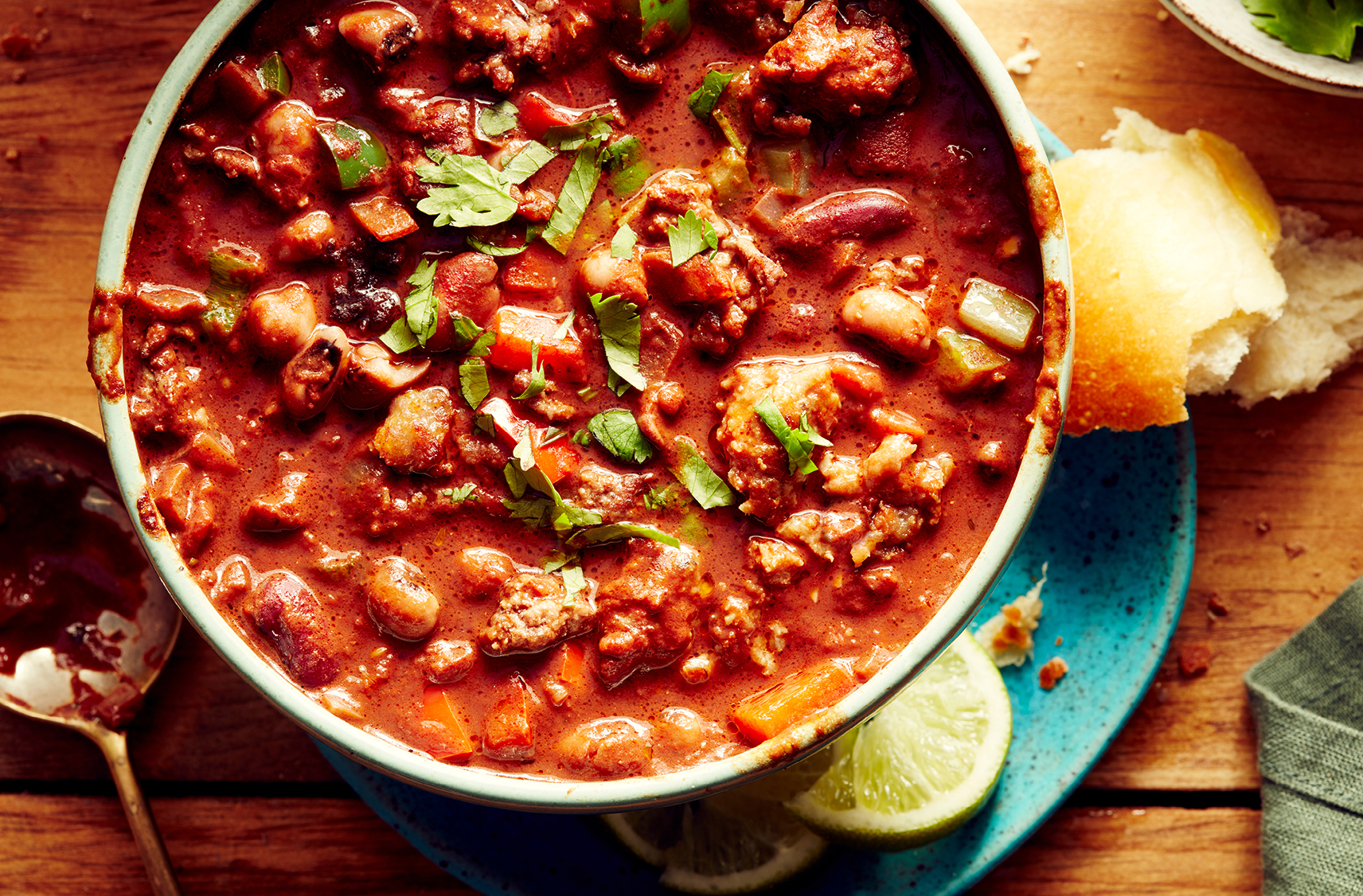 Un chili au bœuf et aux saucisses garni de persil haché servi dans un bol à soupe gris avec des morceaux de pain et des quartiers de lime sur le côté