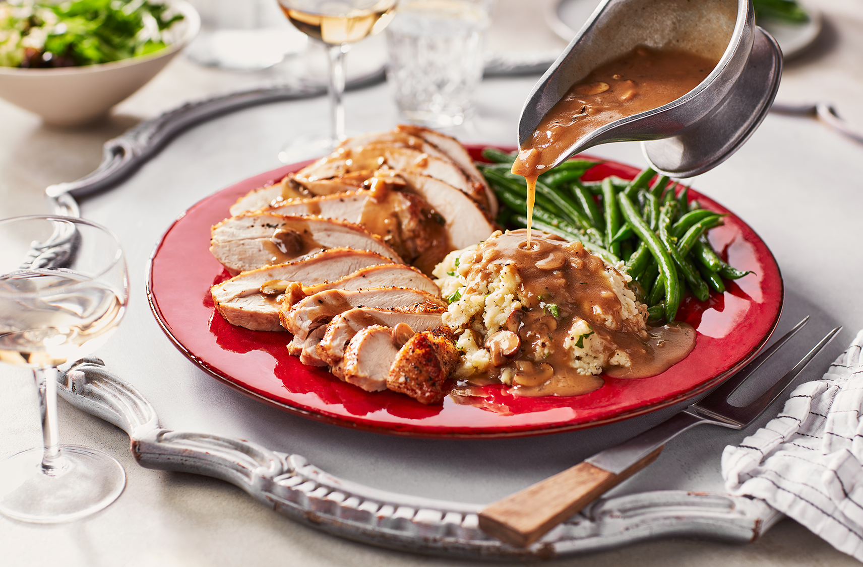 A gravy boat filled with a herbed mushroom chicken bone broth gravy being poured over a plate of turkey stuffing and string beans.