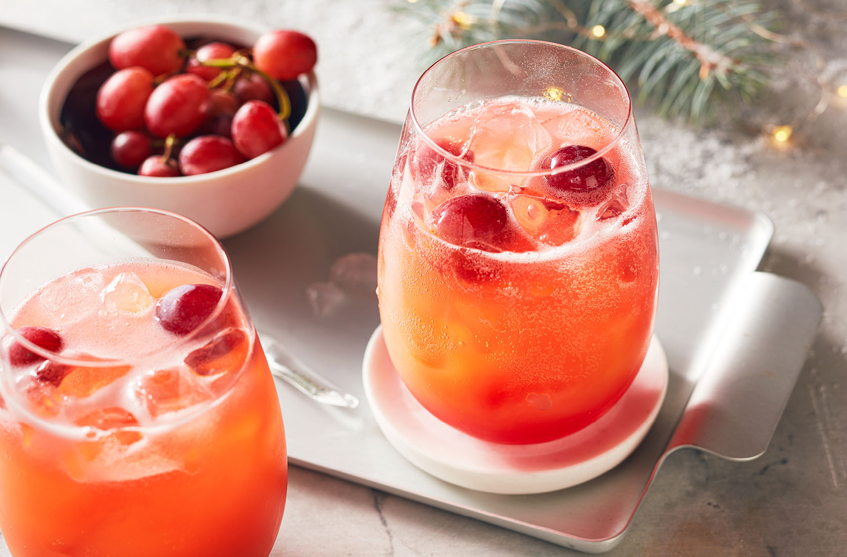 Deux verres de jus pétillant avec des glaçons et des raisins rouges dans un bol sur un plateau de service argent.