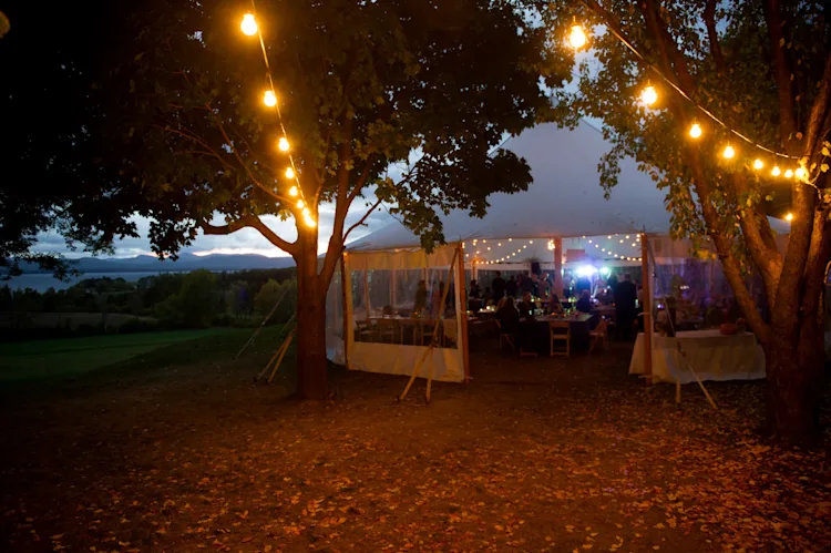 Wedding reception tent glowing with string lights at dusk surrounded by fall foliage with the Adirondack Mountains in the distance