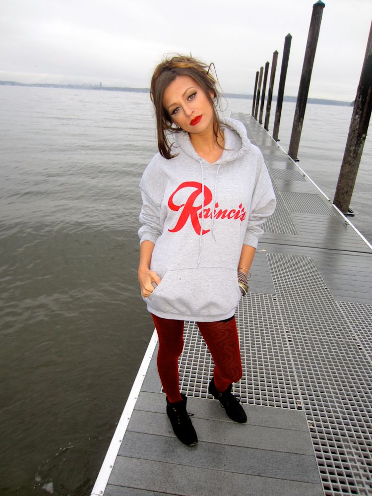 Cassandra McClure standing on a Puget Sound dock in The North West Clothing's grey raincity hoodie and red tights, downtown Seattle skyline in the distance