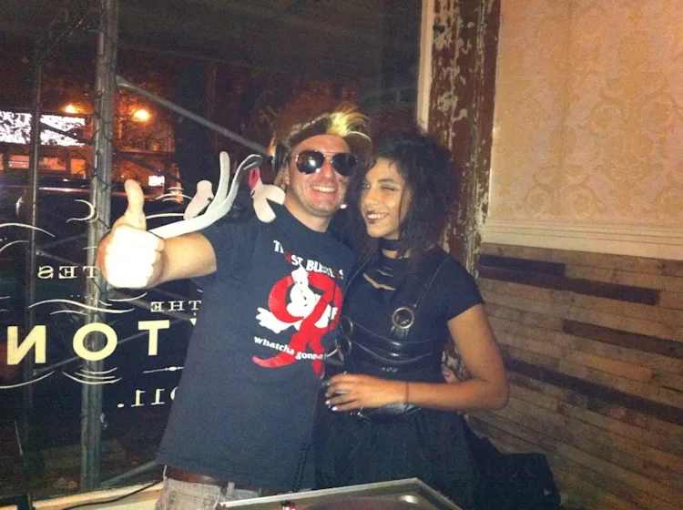 DJ Audeos in a Ghostbusters t-shirt and blonde wig posing with a guest at the Howl-O-Ween Halloween party at The Sexton Bar in Ballard, Seattle