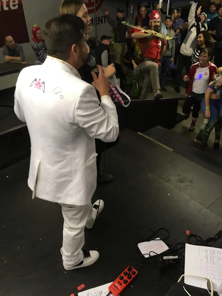 Emcee in a white suit addressing the crowd from the stage during NWAC SnowBash 2016 at evo Seattle