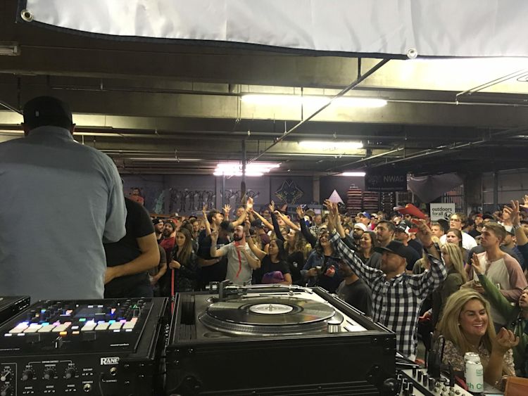 Crowd at NWAC Snowbash with hands in the air, viewed from behind the DJ booth at evo's Fremont flagship, Rane mixer and Technics turntable in the foreground, NWAC logo on the back wall