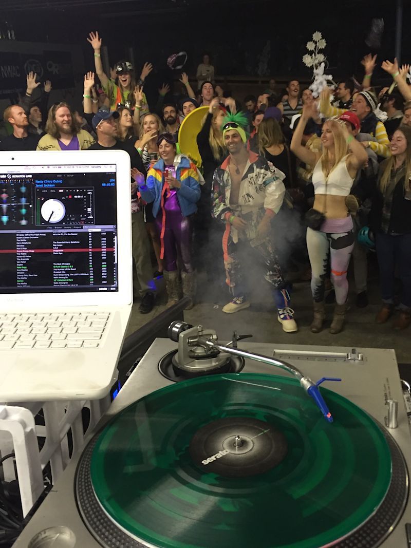View from behind the Serato turntable with green vinyl spinning Janet Jackson while the costumed crowd dances with hands up at NWAC SnowBash 2016 at evo Seattle