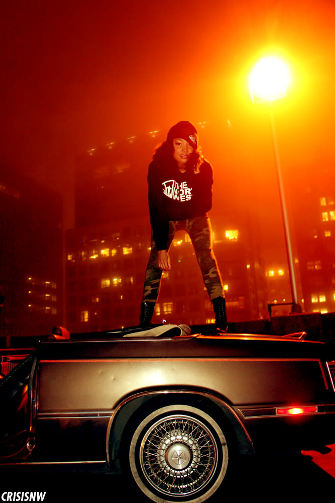 Neko Jamilla standing on a convertible trunk in a The North West hoodie, foggy Seattle skyline behind her