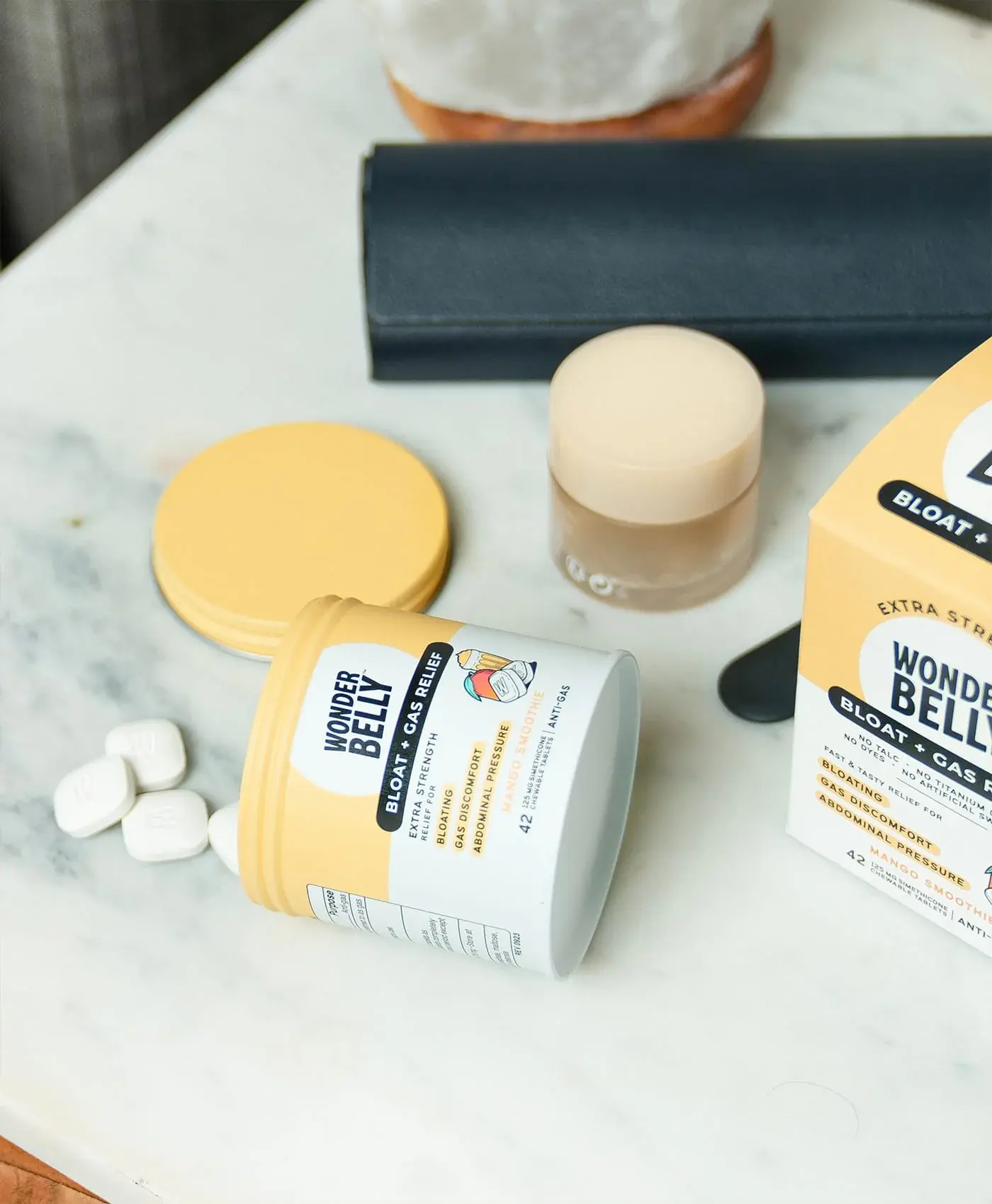 Wonderbelly bloat and gas relief container labeled “Mango Smoothie” lying open on a marble tabletop, with white chewable tablets, product lid, and box nearby in a minimal indoor setting.