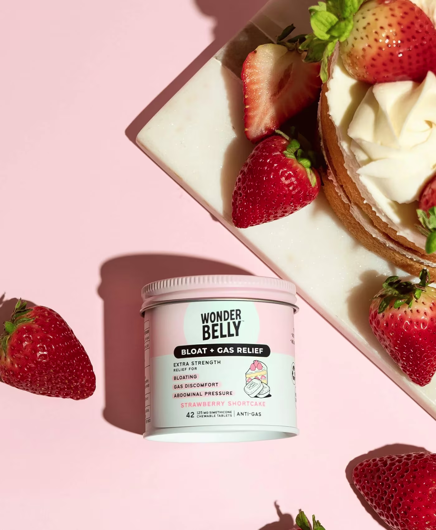Wonderbelly bloat and gas relief jar labeled “Strawberry Shortcake” surrounded by fresh strawberries and a strawberry shortcake dessert on a pale pink background.