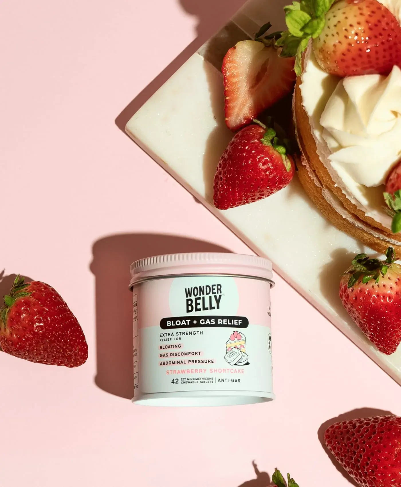 Wonderbelly bloat and gas relief jar labeled “Strawberry Shortcake” surrounded by fresh strawberries and a strawberry shortcake dessert on a pale pink background.